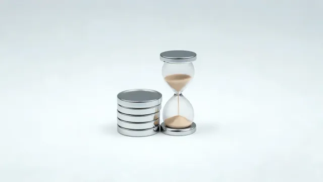 A stack of chrome coins beside a glass hourglass on a light grey background.