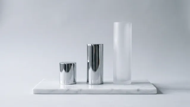 Three cylinders on a white marble slab, growing from short to tall: two in polished chrome, the tallest in frosted glass, on a light grey background.