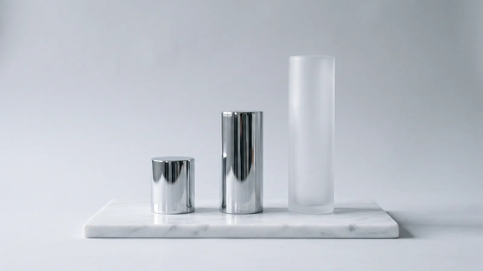 Three cylinders on a white marble slab, growing from short to tall: two in polished chrome, the tallest in frosted glass, on a light grey background.